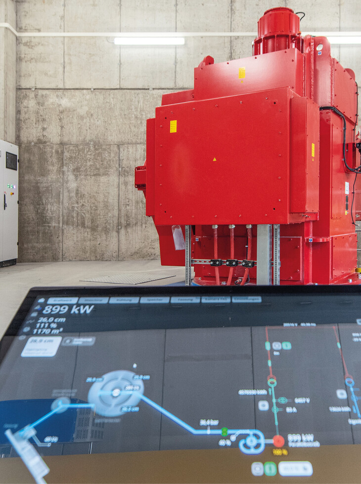 Großes rotes Wasserkraftwerk in einem Betonraum mit einem Bildschirm im Vordergrund, der technische Daten und ein Flussdiagramm anzeigt.