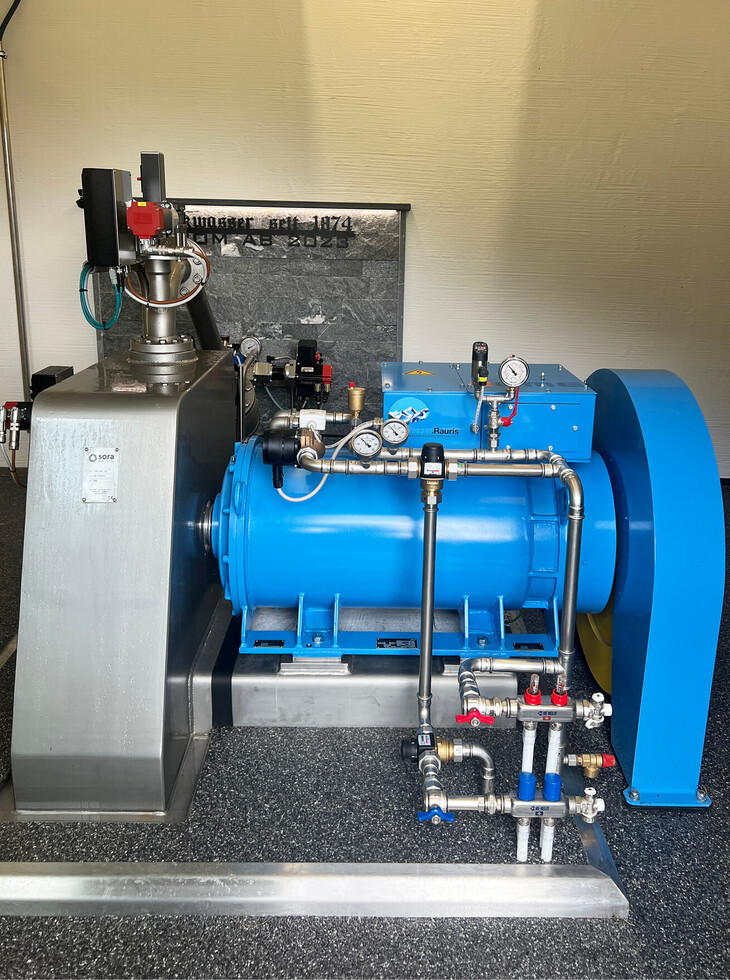 Blue painted drinking water turbine with pressure gauges, pipes and a silver casing in a room with a grey floor.