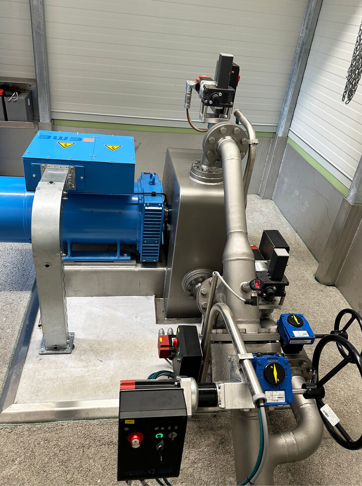 Drinking water turbine with blue electric motor, grey pipes and control unit in a room with white walls.