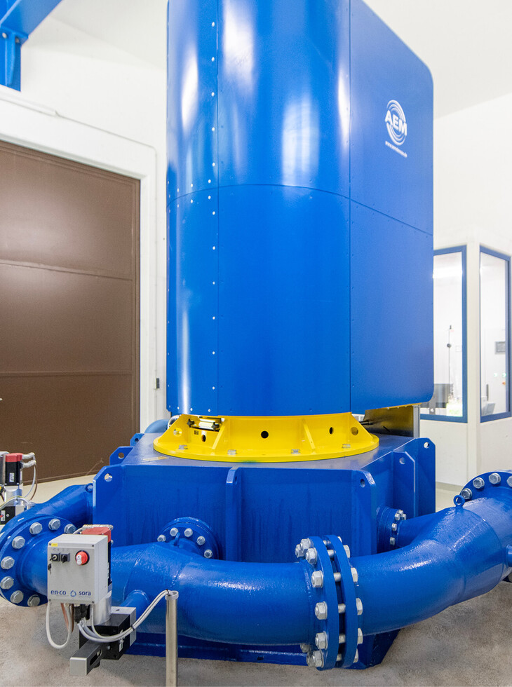 Large blue hydroelectric power plant with yellow base and connected pipes in a bright room.
