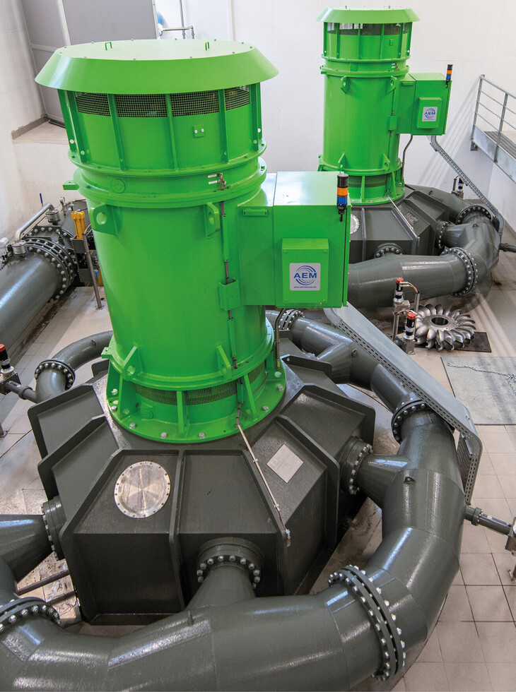 Two green, cylindrical hydroelectric power plants with grey pipes in a bright industrial building.