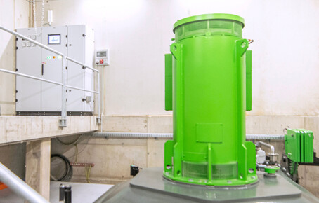 Grün lackiertes Wasserkraftwerk in einem hellen Raum mit Steuerungstafel an der Wand.