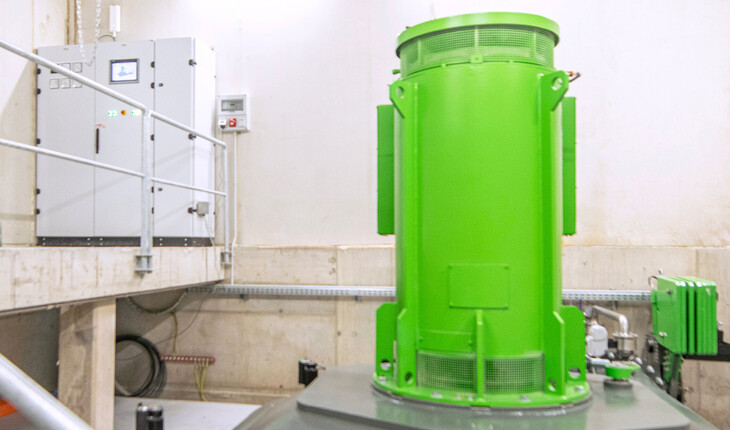 Grün lackiertes Wasserkraftwerk in einem hellen Raum mit Steuerungstafel an der Wand.