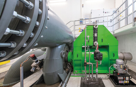 Große graue Rohrleitung mit Flanschverbindung und grünes Wasserkraftwerk mit Messinstrumenten in einem technischen Raum.