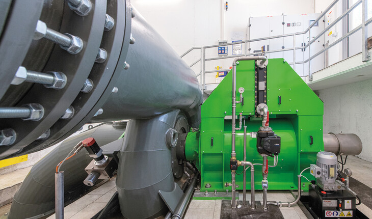 Große graue Rohrleitung mit Flanschverbindung und grünes Wasserkraftwerk mit Messinstrumenten in einem technischen Raum.