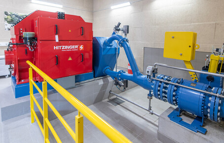 Rotes Wasserkraftwerk mit blauem Rohrleitungssystem in einem technischen Raum mit gelbem Geländer.