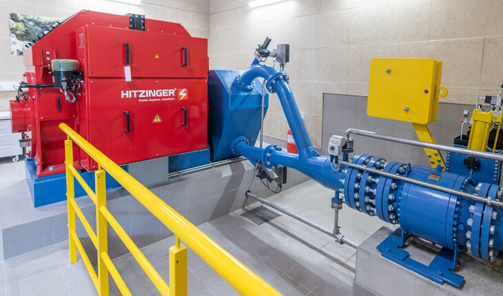 Rotes Wasserkraftwerk mit blauem Rohrleitungssystem in einem technischen Raum mit gelbem Geländer.