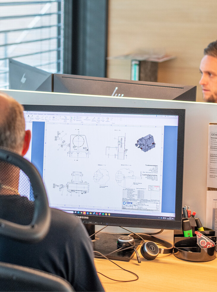 Two people at an office desk with a computer screen displaying technical drawings and various desk utensils.