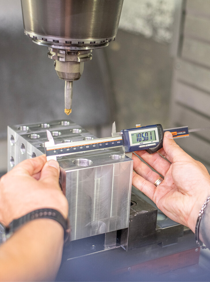 Hands measure a metal workpiece with digital calipers in front of a CNC milling machine.