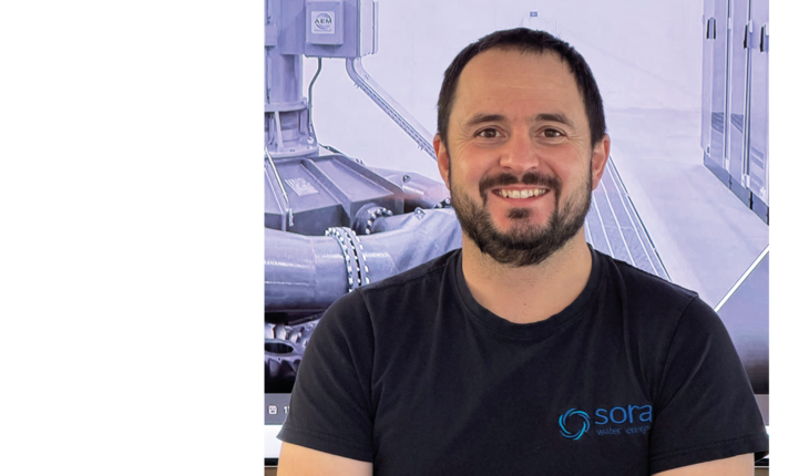 Oberkörper eines Mannes mit Bart, der ein schwarzes T-Shirt mit dem Logo und Schriftzug 