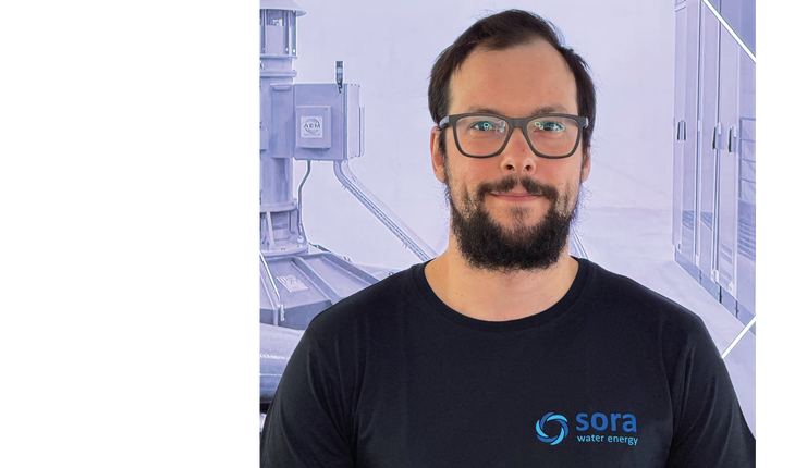 Il torso di un uomo con la barba, che indossa una maglietta nera con il logo e la scritta 