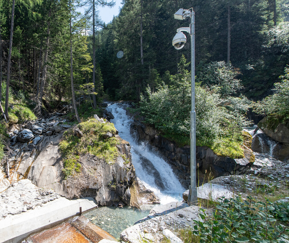 Cascata in una valle boscosa con una telecamera di sorveglianza montata su un palo alto in primo piano.